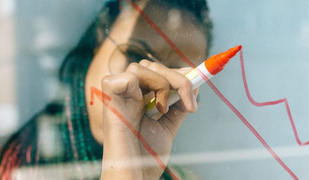 Woman in a Beige Coat Writing on a Glass Panel Using a Whiteboard Marker