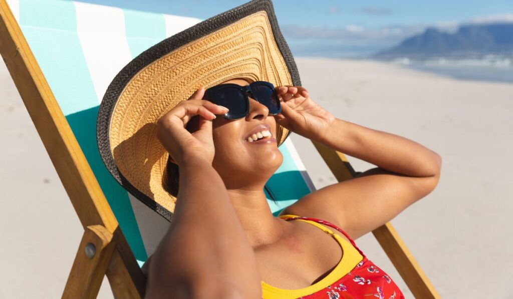 Smiling mixed race woman on beach holiday sitting in deckchair wearing sunglass sunbathing