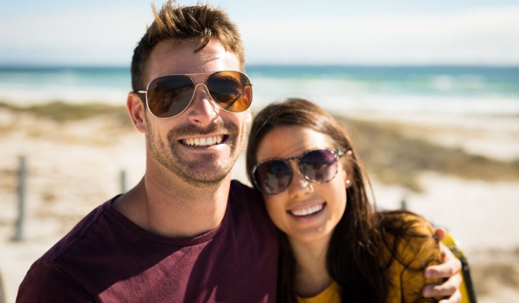 Portrait of happy caucasian couple sitting on beach by the sea embracing wearing sunglasses smiling