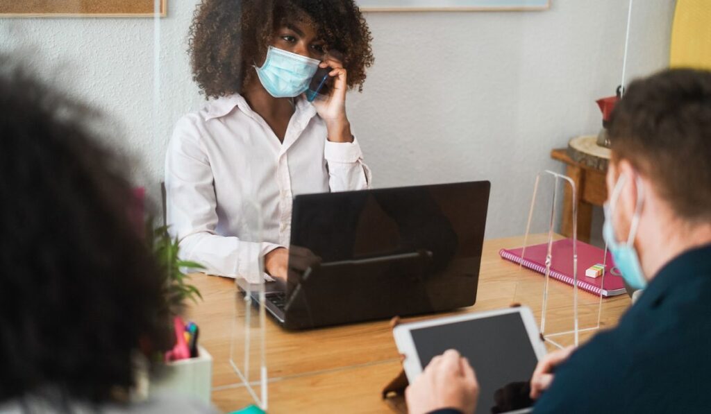 Multiracial coworkers with face masks inside modern office working behind safety plexiglass