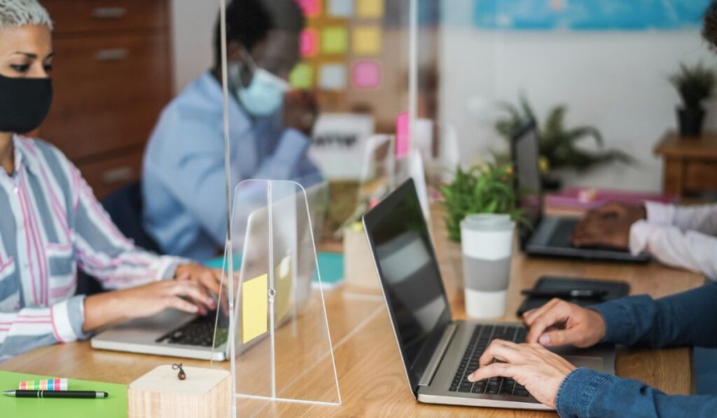 Multiracial coworkers with face masks inside modern office working behind safety plexiglass - Focus on right hands